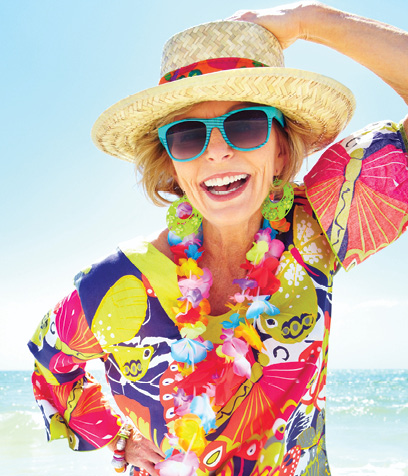 senior blonde woman smiling on the beach in a colorful blouse, sun hat and teal sunglasses